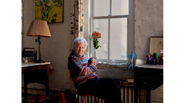 Lois Dodd in her East Village apartment with her painting “Eli In Apple Tree” (1965) above the table.Credit...Vincent Tullo for The New York Times