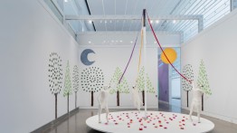 A photo of the Queens Museum's gallery containing an installation with three white, life-sized dog sculptures in mid-frolick on a white, circular platform and holding lavender, red, and orange ribbons attached to a large white cane pole. The platform is covered in pink, magenta, and red paper mache flowers. The gallery walls are covered with trees made of light and dark green painted paper mache leaves. The gallery also features a grey-blue crescent moon and a large orange sun floating above back entrance.